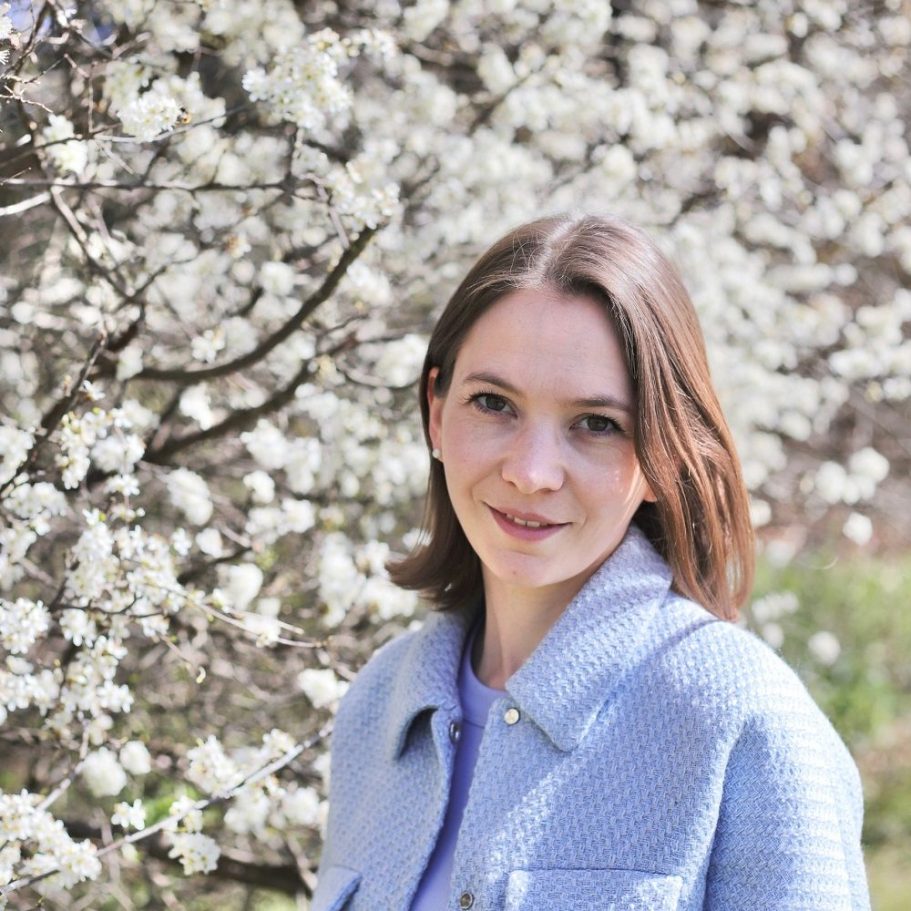 Katharina Huber, Floristik Freelancer, MünchenFlorales Design, Meisterfloristin Junge Frau vor blühenden Bäumen mit weißen Blüten, lächelnd in einem blauen Mantel. Katharina Huber-Florales Design